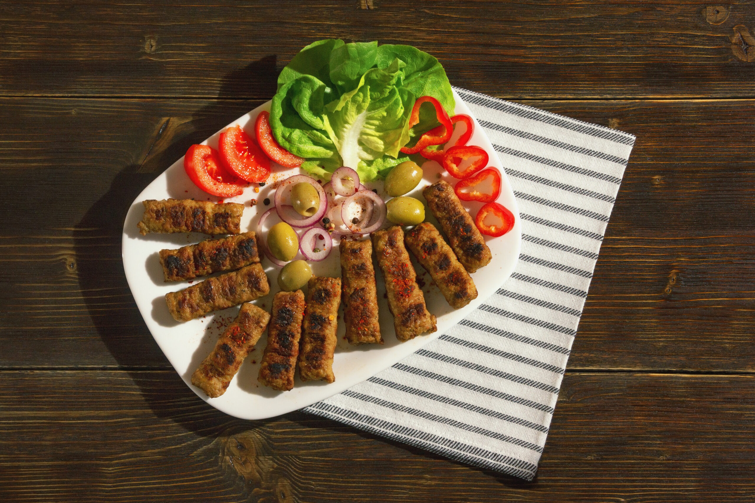 Balkan cuisine. Cevapi - grilled dish of minced meat- with vegetables. Dark rustic background. Flat lay